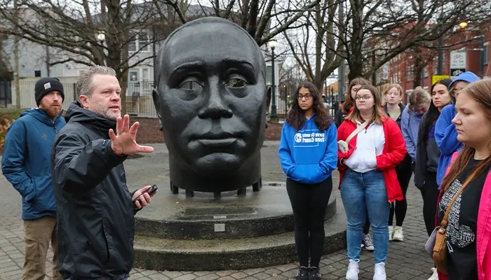 an instructor stands in front of a sculpture and speaks to students